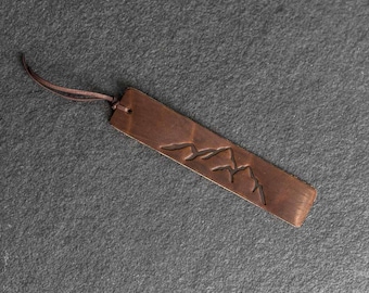 Könnte beinhalten: Ein kupferfarbener Lesezeichen mit einem Bergketten-Design. Das rechteckige Lesezeichen hat oben eine Lederschnur befestigt. Die Bergkette ist in das Metall geätzt. Das Lesezeichen hat eine warme braune Farbe.