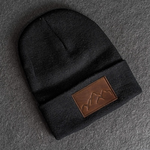 Pode incluir: Gorro preto com um remendo de couro castanho com um desenho de cordilheira. O gorro é feito de um material de malha e tem uma aba dobrada. O remendo é retangular e costurado na frente.