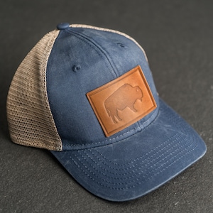 May include: A blue and brown trucker hat with a leather patch featuring a silhouette of a buffalo. The hat has a mesh back.