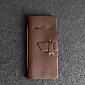 May include: A brown leather Hunting Log journal with a strap closure. The journal has the words "HUNTING LOG" embossed on the cover, along with a deer silhouette. The journal is closed with a leather strap and two brass rivets.