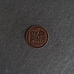 May include: A round, brown leather patch with the embossed text "OX & PINE LEATHER GOODS CO." and a bull silhouette. The patch is circular and has a slightly raised border, set against a dark gray background.