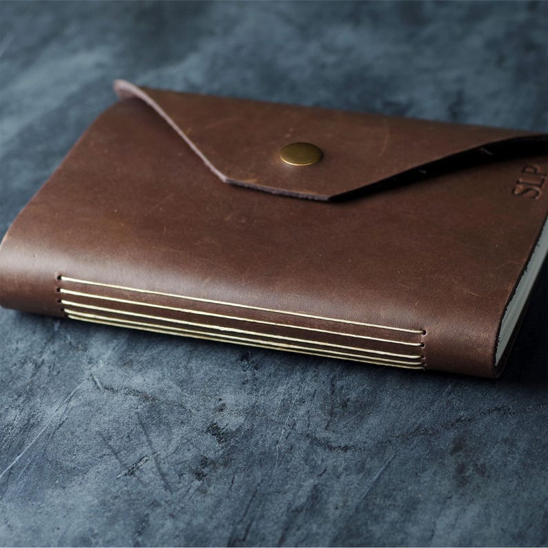 May include: A dark brown leather journal with a triangular envelope-style flap secured by a single brass-colored metal snap button. The spine features visible cream-colored thread stitching, and the letters SLP are embossed on the front cover.