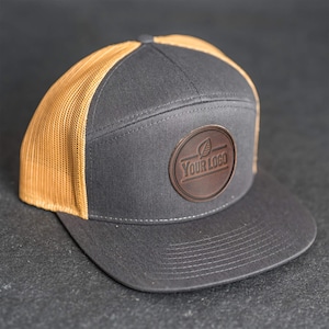 Puede incluir: Gorra de camionero gris carbón con un parche de cuero marrón. El parche presenta un diseño de hoja y el texto "YOUR LOGO". La gorra tiene una visera curva y una parte trasera de malla color canela.