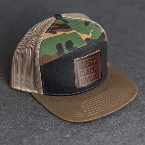Puede incluir: Gorra de camionero con visera marrón y panel frontal negro. El panel superior presenta un patrón de camuflaje. Un parche de cuero marrón en la parte delantera dice "BEST DAD EVER."