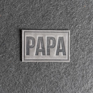 Puede incluir: Un parche de cuero gris con la palabra "PAPA" en relieve en letras negras.