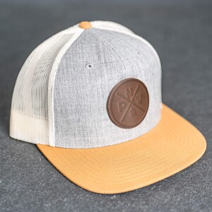 May include: A grey and orange trucker cap with a brown leather patch. The cap has a white mesh back, a curved orange peak, and a brown button on top. The patch features a circular design with the letters "N", "W", and "P" and a tree.