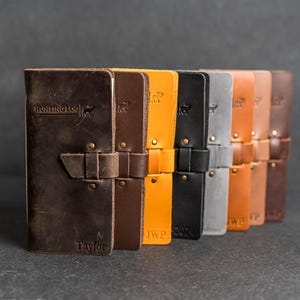 May include: A collection of leather journals in various colors, including brown, orange, black, and gray. Each journal features a strap closure and is embossed with text and a deer silhouette. The first journal reads "HUNTING LOG" and "Taylor".