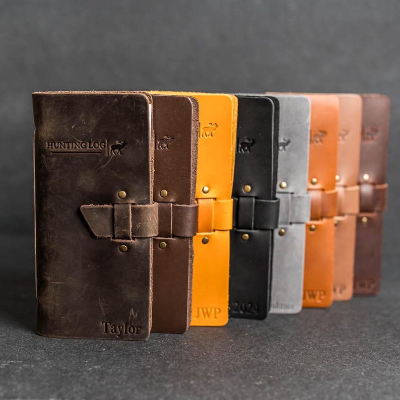 May include: A collection of leather journals in various colors, including brown, orange, black, and gray. Each journal features a strap closure and is embossed with text and a deer silhouette. The first journal reads "HUNTING LOG" and "Taylor".