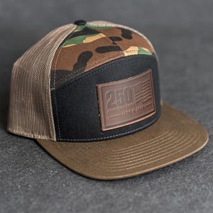 Peut inclure: Casquette de camionneur marron et noire avec un motif camouflage sur le dessus. Le devant noir porte un écusson en cuir marron avec l'inscription "250 ANNIVERSAIRE 1776-2026" et un drapeau américain. L'arrière est en filet.