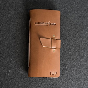 May include: A brown leather fishing log with the words "FISHING LOG" embossed on the front. The log has a strap closure with brass rivets and the initials "JWP" are stamped at the bottom.