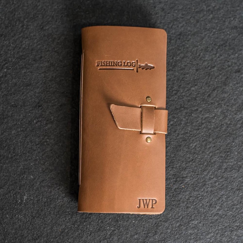 May include: A brown leather fishing log with the words "FISHING LOG" embossed on the front. The log has a strap closure with brass rivets and the initials "JWP" are stamped at the bottom.