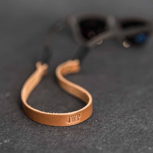 May include: A light brown leather eyeglass strap with the initials "JWP" embossed on the strap. The strap is connected to a pair of black sunglasses. The strap is designed to be worn around the neck to keep glasses secure.