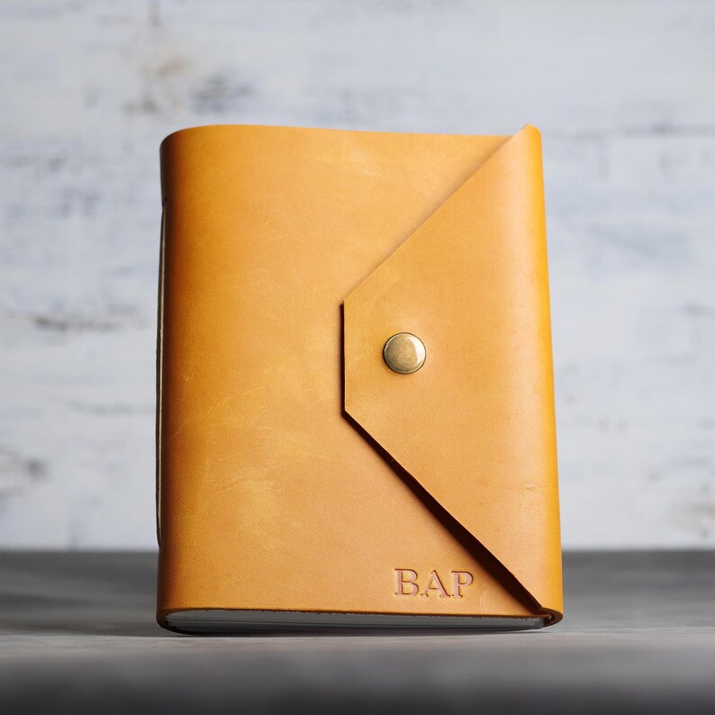 May include: A tan leather journal with a triangular flap closure secured by a single brass-colored metal snap button. The front cover features the embossed letters B.A.P. in the bottom right corner, showcasing a smooth, matte finish.