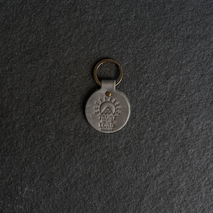 Op de afbeelding: Een grijze leren sleutelhanger met een messing ring. De ronde hanger heeft de reliëftekst "TRUST IN THE LORD" boven een berg- en zonontwerp. De achtergrond is een donkergrijze, getextureerde ondergrond.