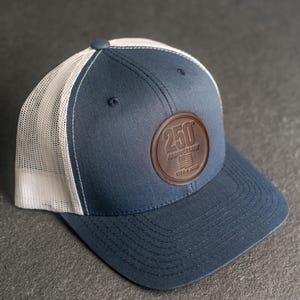 以下が含まれることがあります： ネイビーブルーのメッシュバックトラックキャップ。茶色の革製パッチには「250 ANNIVERSARY」と「1776-2026」の年号が記されています。カーブしたつばとトップにボタンが付いています。