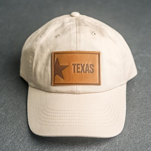 May include: A beige baseball cap featuring a rectangular tan leather patch centred on the front. The patch is embossed with a five-pointed star on the left side and the word TEXAS in capital letters on the right side, with stitched detailing.