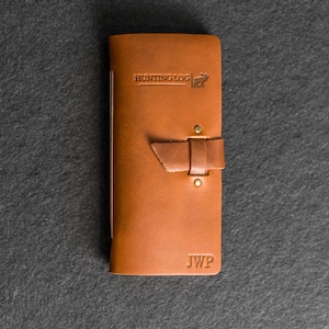 May include: A brown leather hunting log with the text "HUNTING LOG" and a deer graphic embossed on the cover. The log has a strap closure and the initials "JWP" embossed on the bottom.