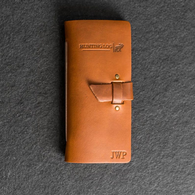 May include: A brown leather hunting log with the text "HUNTING LOG" and a deer graphic embossed on the cover. The log has a strap closure and the initials "JWP" embossed on the bottom.