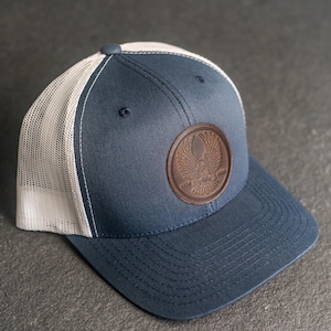 May include: Navy blue trucker hat with a white mesh back and a curved brim. The hat features a brown leather patch with an embossed eagle design and the numbers 1770 and 2016.