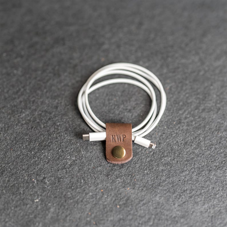 May include: A white charging cable coiled and secured with a brown leather cord organizer. The leather strap features the embossed letters KWP and a circular brass-toned metal snap button fastener to keep the cable neatly bundled together.