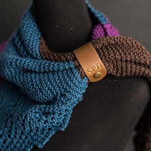 Op de afbeelding: Een gebreide sjaal met secties van blauwgroen, bruin en paars garen. Een beige leren label is bevestigd met een messingkleurige metalen drukknoop. Het leren label is voorzien van de hoofdletters SPB in een duidelijk lettertype.