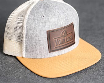 Könnte beinhalten: Grau-gelbe Baseballkappe mit Mesh-Rückseite. Auf der Vorderseite befindet sich ein brauner Lederaufnäher mit dem Text "YOUR LOGO" und einem Blattdesign. Der Schirm ist gelb und der obere Knopf ist hellbraun.