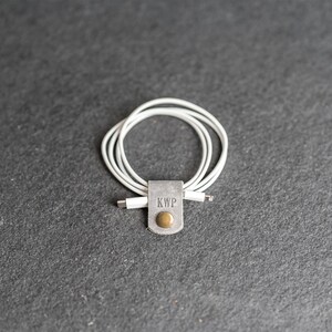 May include: A white charging cable coiled and secured with a grey leather strap. The strap features the embossed letters KWP and a circular brass-colored metal snap button fastener centered on the front of the leather tab.