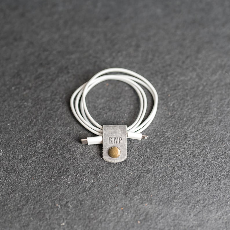 May include: A white charging cable coiled and secured with a grey leather strap. The strap features the embossed letters KWP and a circular brass-colored metal snap button fastener centered on the front of the leather tab.