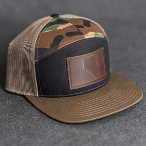 Puede incluir: Gorra de camionero con visera marrón y panel frontal negro con parche de cuero marrón con contorno de estado. La parte superior de la gorra tiene un estampado de camuflaje y los laterales son de malla beige.