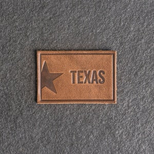Op de afbeelding: Een bruin leren patch met een vijfpuntige ster en het woord "TEXAS" in reliëf in het midden.