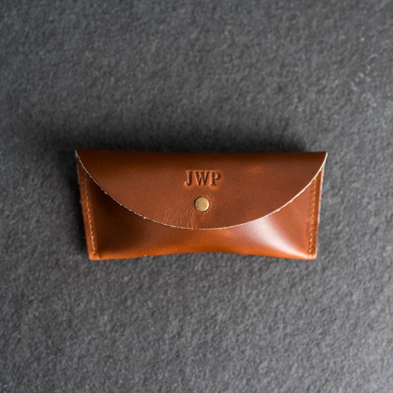 Puede incluir: Un estuche de gafas de cuero marr&oacute;n con cierre de solapa y un broche de lat&oacute;n. Las iniciales "JWP" est&aacute;n grabadas en la parte delantera. El estuche es rectangular y parece hecho a mano.