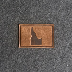 Puede incluir: Un parche de cuero marrón con el estado de Idaho estampado en el centro.