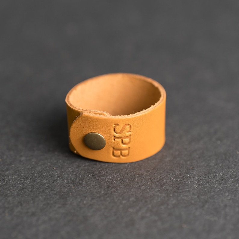 Op de afbeelding: Een beige leren manchetarmband met een ronde koperen drukknoopsluiting en de in reli&euml;f aangebrachte letters SPB op de voorzijde. De band is gemaakt van een enkel stuk glad, lichtbruin leer met zichtbare randen en een eenvoudig ontwerp.