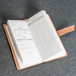 May include: Open leather journal with lined pages and a tab closure. The journal is open to a page with a table labeled "Fish Over 12" and sections for weather and moon phases. The journal is tan and sits on a dark gray surface.