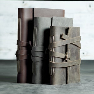 May include: Four leather journals in varying shades of brown and gray. The journals are bound with visible stitching and secured with leather straps. The journals are standing upright, showcasing their textured covers and unique design.