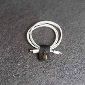 May include: A white charging cable coiled and secured with a black leather cord organizer. The organizer features a brass-colored metal snap button and the embossed letters KWP on the front surface, holding the cable in a neat circular loop.