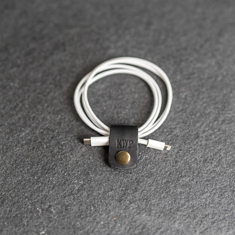 May include: A white charging cable coiled and secured with a black leather cord organizer. The organizer features a brass-colored metal snap button and the embossed letters KWP on the front surface, holding the cable in a neat circular loop.