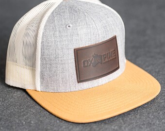 Puede incluir: Gorra de camionero gris y blanca con parche de cuero marrón. La gorra tiene una visera amarilla y un parche de cuero marrón con el texto "OX & PINE". Cierre trasero ajustable.