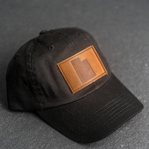 Könnte beinhalten: Eine schwarze Baseballkappe mit einem braunen Lederpatch, auf dem ein stilisierter Buchstabe "I" auf der Vorderseite zu sehen ist.