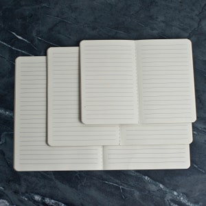 May include: Three lined notebooks of different sizes stacked on top of each other. The notebooks are all closed and have white covers.