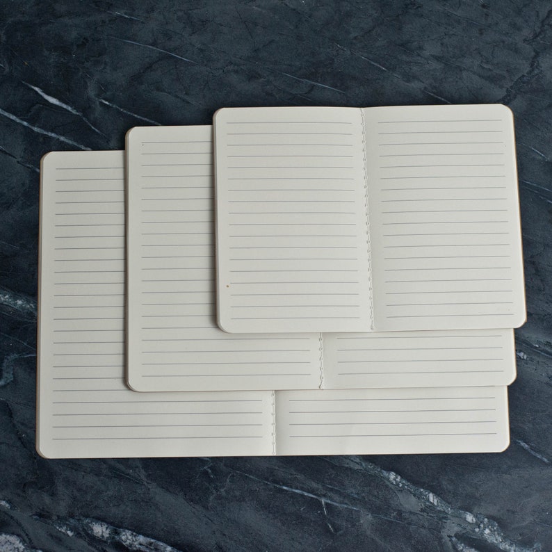 May include: Three lined notebooks of different sizes stacked on top of each other. The notebooks are all closed and have white covers.