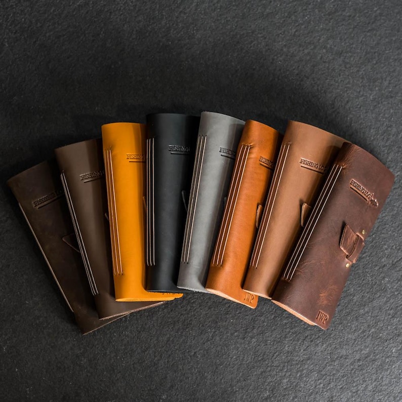 May include: A collection of leather journals in various colors, including brown, orange, black, and gray. Each journal has a vertical design with a strap and the text "FISHING LOG" embossed on the cover. The journals are arranged in a fan shape.