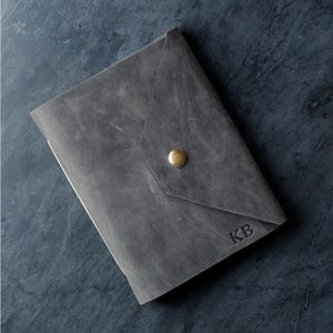 May include: A grey leather journal cover with a triangular flap closure secured by a single brass-colored metal snap button. The front surface features the embossed letters KB in the bottom right corner, showcasing a matte, textured finish.