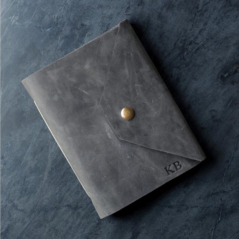 May include: A grey leather journal cover with a triangular flap closure secured by a single brass-colored metal snap button. The front surface features the embossed letters KB in the bottom right corner, showcasing a matte, textured finish.