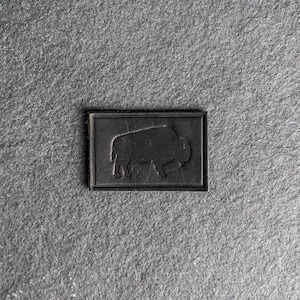 May include: A black leather patch with a silhouette of a buffalo in the center.