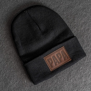 May include: Black knit beanie with a brown leather patch that reads "PAPA" in capital letters. The beanie is folded at the brim and is made of a soft, warm material. A stylish and comfortable hat.