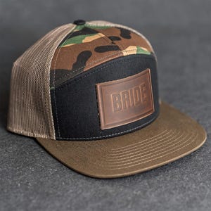 May include: A trucker hat with a camouflage pattern on the top, a black front panel, and a brown brim. The hat features a brown leather patch with the word "BRIDE" embossed on it. The mesh back is tan.