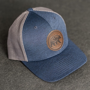 May include: A blue baseball cap with a grey mesh back. The cap has a brown leather patch with a bear and mountain design. The hat is a trucker style.