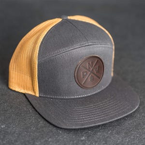 Op de afbeelding: Een grijze en bruine truckerpet met een platte klep. Het voorpaneel is grijs met een bruine leren patch met de letters "PNW" en een boomontwerp. De mesh achterkant is bruin.