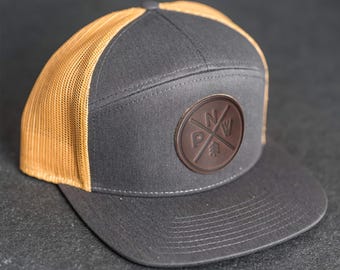 Puede incluir: Gorra de camionero gris y beige con visera plana. El panel frontal es gris con un parche de cuero marrón con las letras "PNW" y un diseño de árbol. La parte trasera de malla es beige.